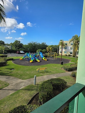 A playground with slides and a bench, surrounded by well-maintained grass and palm trees. - Beach-5 Pools-25 Min from airport-Unit with reserve water system  (Loíza)