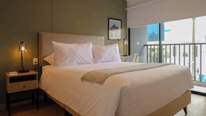 Cama bem arrumada com cabeceira, mesa de cabeceira com abajur, e janela com vista para prédios.