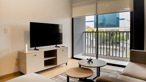 Una habitación de hotel moderna con un televisor grande sobre un mueble de madera, una mesa redonda, un sofá y un balcón con vistas a la calle y un edificio.