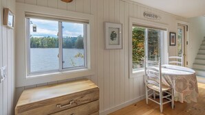 A room with a large window overlooking a lake, a wooden bench, a dining table with chairs, and a staircase leading to another level. - Pondside Cottage w/ Dock & Patio! [Loons Nest] (Mount Desert)