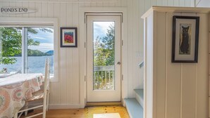 A room with a view of a lake, a door leading outside, a table with a patterned tablecloth, and two framed pictures on the wall. - Pondside Cottage w/ Dock & Patio! [Loons Nest] (Mount Desert)