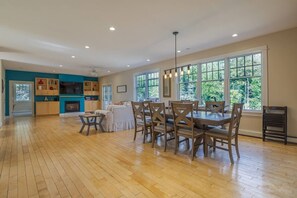 A spacious living area with a dining table and chairs, a fireplace, and large windows offering a view of greenery. - Spacious 5BR Home in Center of MDI [Loons Landing] (Mount Desert)
