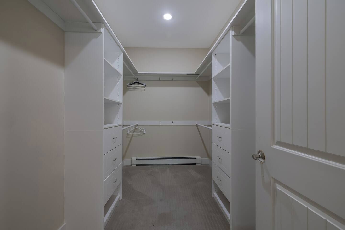 A narrow walk-through closet with white shelving, a radiator, and a door on the right.