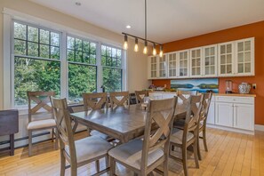 A dining area with a large table and chairs, a kitchen with white cabinets, and a view of trees through large windows. - Spacious 5BR Home in Center of MDI [Loons Landing] (Mount Desert)