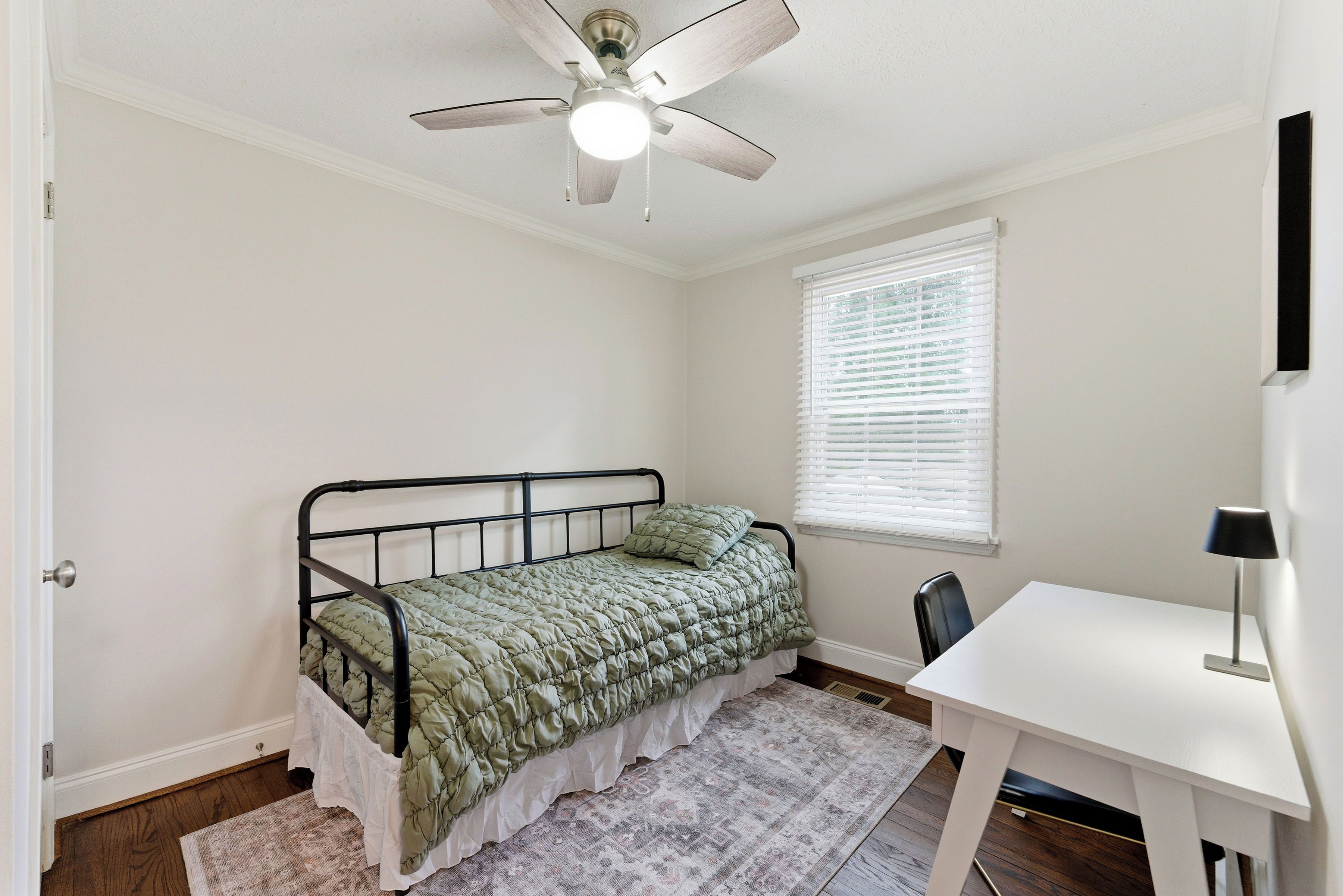 A bedroom with a single bed, a desk, a chair, a ceiling fan, and a window with blinds.