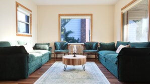 Ein Wohnzimmer mit einem grünen Sofagarnitur, einem runden Couchtisch und Blick durch ein großes Fenster.