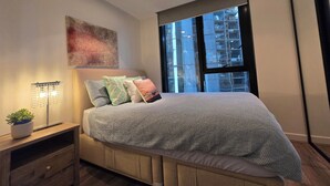 Ein modernes Schlafzimmer mit einem großen Bett, einem Nachttisch mit Lampe und Blick auf ein Gebäude durch ein großes Fenster.