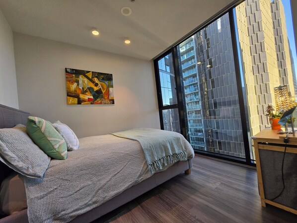 Ein modernes Schlafzimmer mit großem Fenster, einem Bett mit gemusterten Betttextilien, einem Bild an der Wand und Blick auf ein hohes Gebäude.