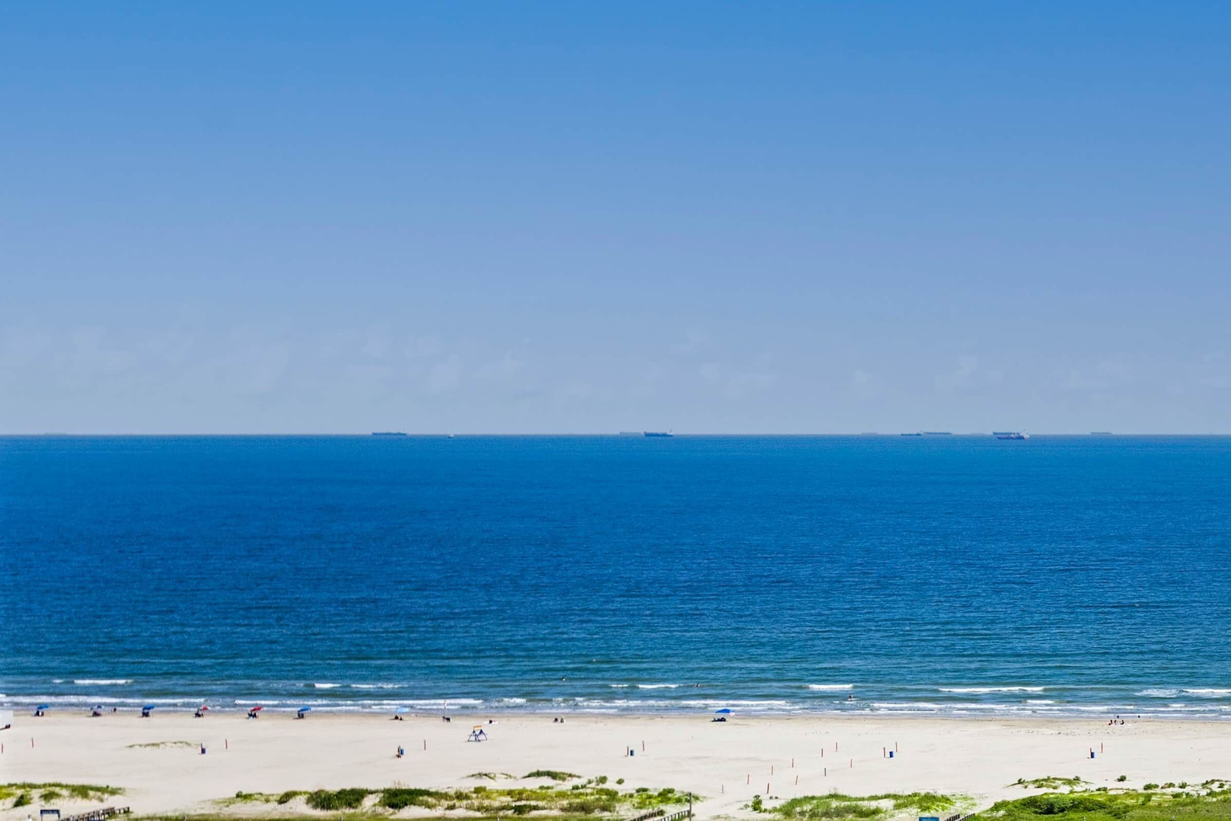 Una playa con cielo despejado, mar tranquilo y una orilla arenosa con gente y sombrillas.