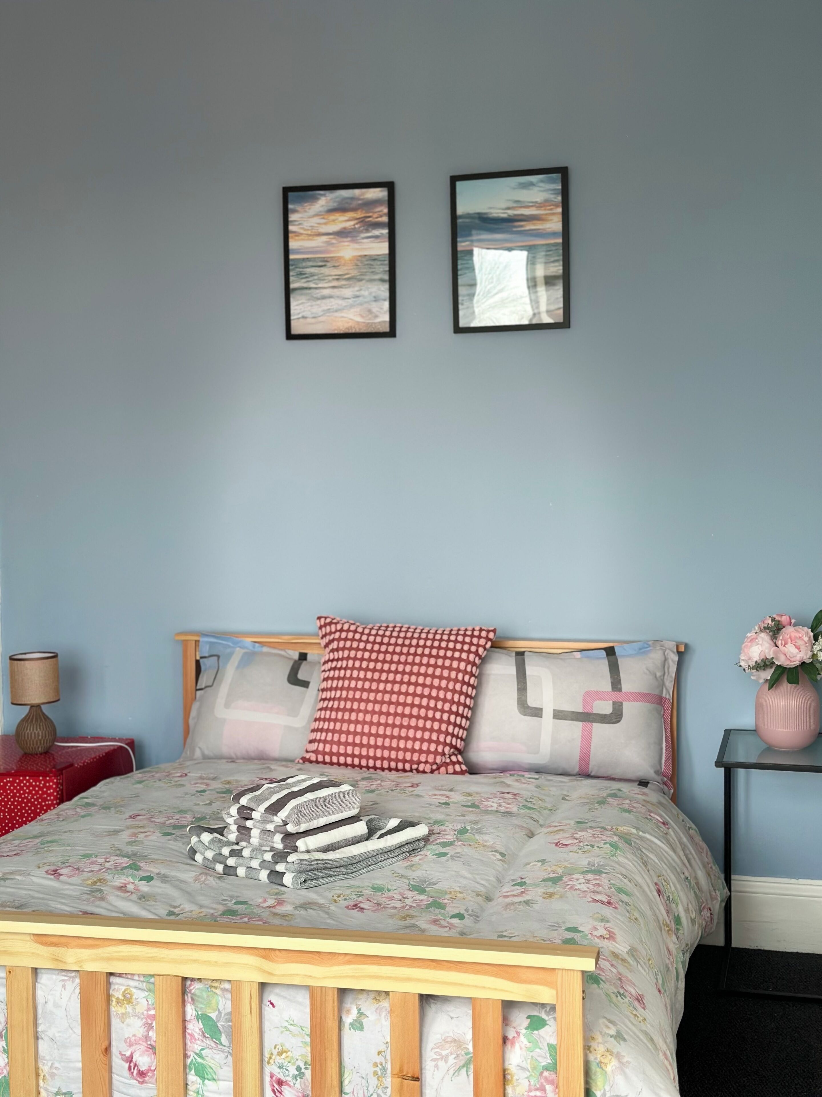 A bedroom with a wooden bed, floral bedding, a red and white patterned pillow, and two framed pictures on the wall.
