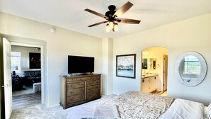 Slaapkamer met een bed, een dresser, een plafondventilator, een televisie, een schilderij, een spiegel in de badkamer en een deur naar een andere kamer.