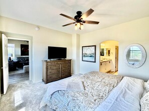A bedroom with a bed, a dresser, a ceiling fan, a TV, a painting, a bathroom mirror, and a door leading to another room. - Tucson Mountain & Wildlife Views in Vistoso (Oro Valley)