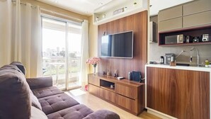 Un salon moderne avec un canapé, un téléviseur fixé sur un mur en bois et un coin cuisine équipé d’appareils électroménagers.