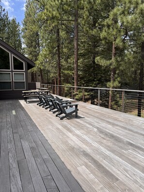 Een houten terras met Adirondack-stoelen en een leuning, omringd door hoge dennenbomen.