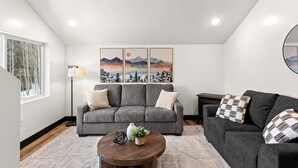 Uma sala de estar moderna com um sofá cinzento, uma mesa de centro redonda e um quadro de montanhas.