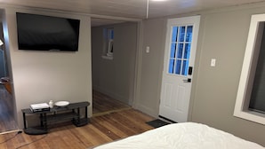 A bedroom with a bed, a TV mounted on the wall, a small table with a bowl and a book, and a door with a window. - Cozy Private Studio w/ Kitchenette (Pittsburgh)