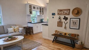 Ein modernes Wohnzimmer mit einer Couch, einem Couchtisch und einer Bank mit Zierkissen. Im Hintergrund ist eine Küche mit weißen Schränken und einem Fenster erkennbar.