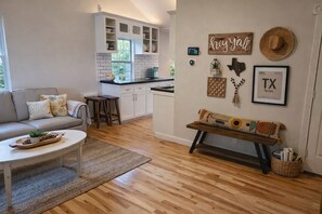 A modern living room with a sofa, coffee table, and a bench with decorative pillows. A kitchen with white cabinets and a window is visible in the background. - Entire Home in League City near NASA & Kemah, Fire Pit, Large Yard, KAYAKS! (League City)