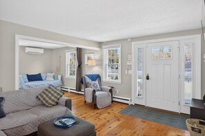 Un salon confortable avec un canapé, un fauteuil et une petite table sur laquelle se trouve un plateau. On y trouve une porte avec une fenêtre, un climatiseur fixé au mur et une lampe.