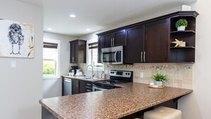 A modern kitchen with dark wooden cabinets, a granite countertop, stainless steel appliances, and a small island with bar stools. - Cottage at Oceanside Village Resort - Sandpiper (Parksville)