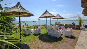 Een terras aan zee met witte rieten meubels, rieten parasols en uitzicht op de zee.