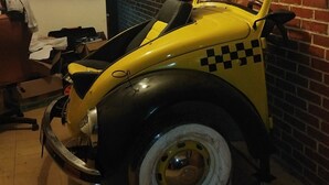 En gul och svart veteranbil med hjul i rutmönster står parkerad i ett rum med en tegelvägg och en klocka.