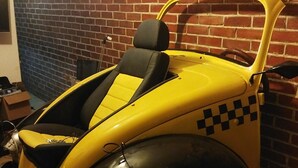 En gammal, gul och svart bil står parkerad inomhus, lutad mot en tegelvägg.