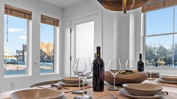 Mesa de jantar posta com tigelas, copos de vinho e garrafas, com vista para a cidade através de grandes janelas.