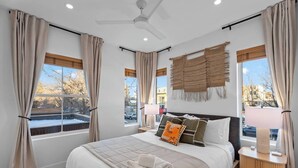 Quarto com cama, mesa de cabeceira, abajur, ventilador de teto e janelas amplas com cortinas.