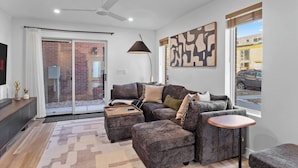 Uma sala de estar moderna com um sofá cinzento, uma pequena mesa de madeira, um abajur e uma grande janela com vista para a rua.