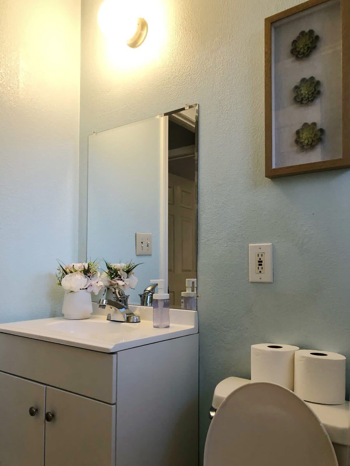 Une salle de bain avec un lavabo blanc, un miroir et des toilettes. Un tableau encadré est accroché au mur et un luminaire se trouve au-dessus du miroir.