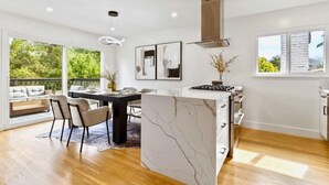 Uma cozinha moderna com uma ilha de mármore, uma área de refeições com uma mesa e cadeiras pretas, e uma janela grande com vista para o verde.