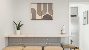 Una cocina con encimera blanca, armarios grises y tres taburetes de bar. Hay una planta en la encimera y una obra de arte enmarcada sobre la encimera.