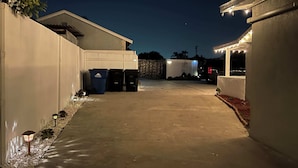 Une allée pavée mène à un bâtiment à l’extérieur blanc, doté d’une porte de garage. Trois poubelles et une guirlande lumineuse sont disposées le long de l’allée.