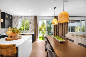 Une cuisine moderne avec un revêtement mural en marbre, un îlot central agrémenté d’un panier de fruits et un coin repas avec une table et des chaises en bois.