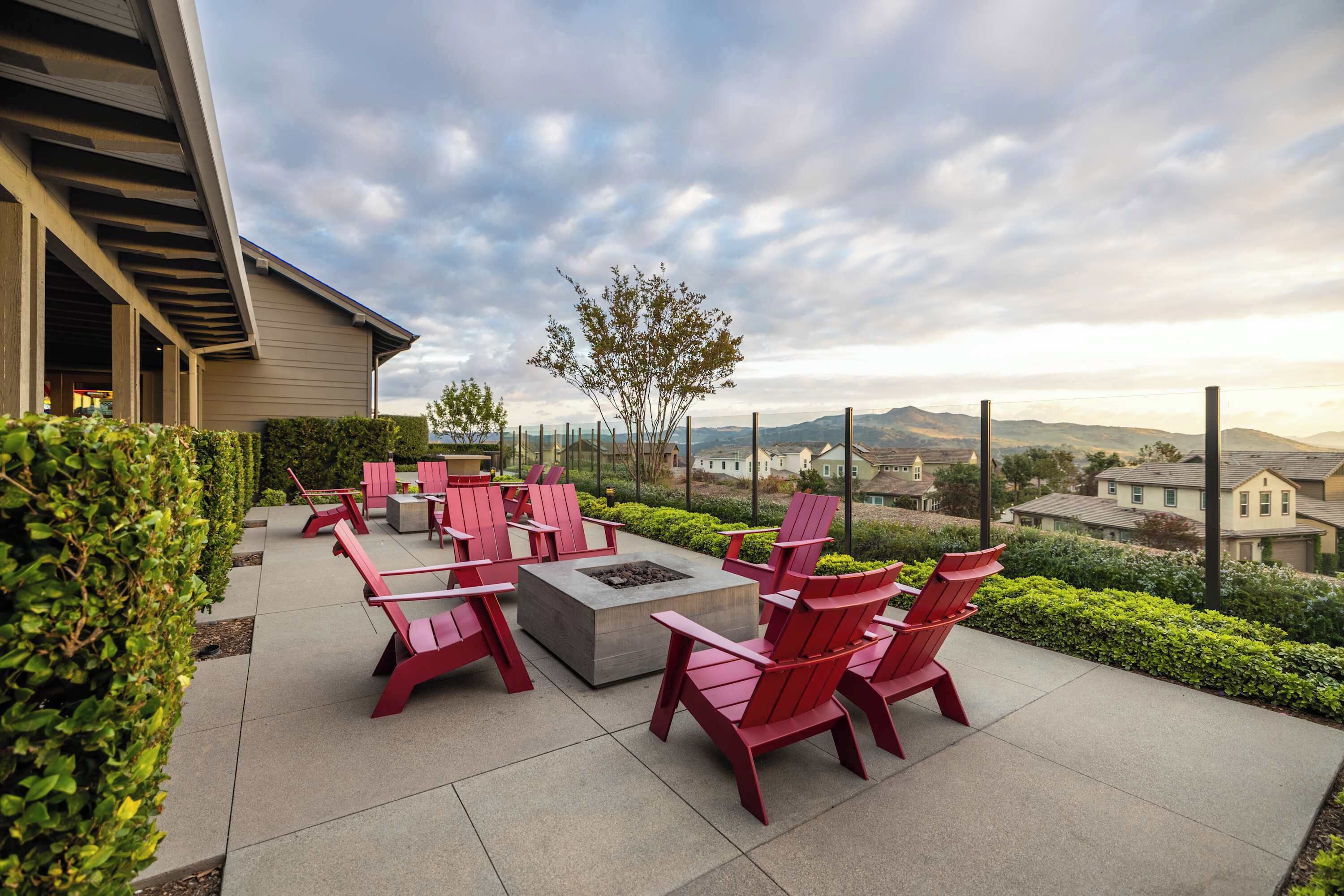An outdoor patio with red Adirondack chairs and a fire pit, overlooking a residential area.