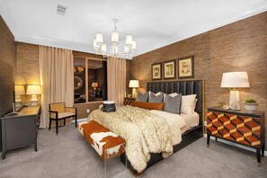 Une chambre d’hôtel moderne avec un grand lit, un bureau et une chaise. Un lustre est suspendu au-dessus du lit, et il y a une commode avec des tiroirs.