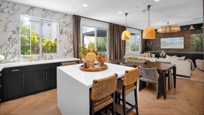 A modern kitchen with a central island, dark cabinetry, and a dining area with wicker chairs and a wooden table. - Marbella Lane - Stylish Dream Home in RMV w Pool (Mission Viejo)