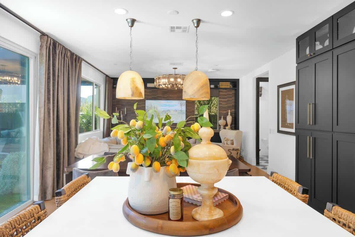 Un comedor moderno con una mesa blanca, sillas de mimbre, un florero con flores amarillas y una bandeja de madera.