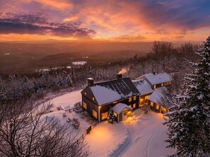 Una casa cubierta de nieve con una chimenea grande, rodeada de árboles y un paisaje nevado.