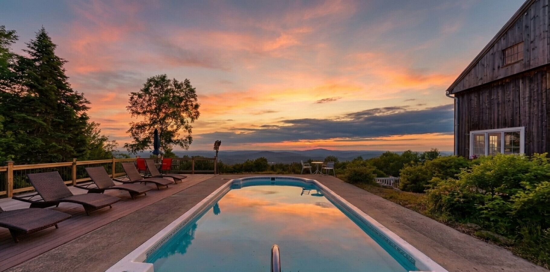Une piscine rectangulaire, dotée de chaises longues et d’une terrasse en bois, entourée de verdure et offrant une vue sur des montagnes lointaines.