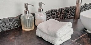 Une salle de bain avec un mur en pierre, un lavabo blanc et deux distributeurs de savon transparents.