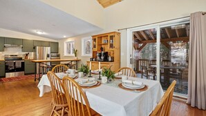 A dining area with a table set for four, wooden chairs, a kitchen in the background, and a view through a glass door. - Bear Den: Hot Tub + Secluded | Sleeps 8 | Modern (Wardsboro)