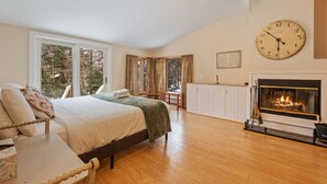Ein geräumiges Schlafzimmer mit einem großen Bett, einem Kamin, einer Uhr und Blick auf Bäume durch das Fenster.