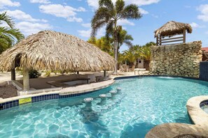 A clear swimming pool with a thatched-roof structure and stone walls, surrounded by palm trees. - Boat House with pool and free parking (Kralendijk)