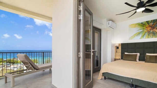 A balcony with a view of the ocean, a bed with a green headboard, and a ceiling fan. - Bonaire Beach Apartments 14 with spectacular sea views and beautiful sunsets every evening (Kralendijk)