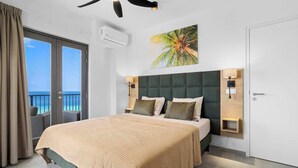 A modern hotel room with a large bed, a balcony with a view, a ceiling fan, and a tropical-themed wall art. - Bonaire Beach Apartments 14 with spectacular sea views and beautiful sunsets every evening (Kralendijk)
