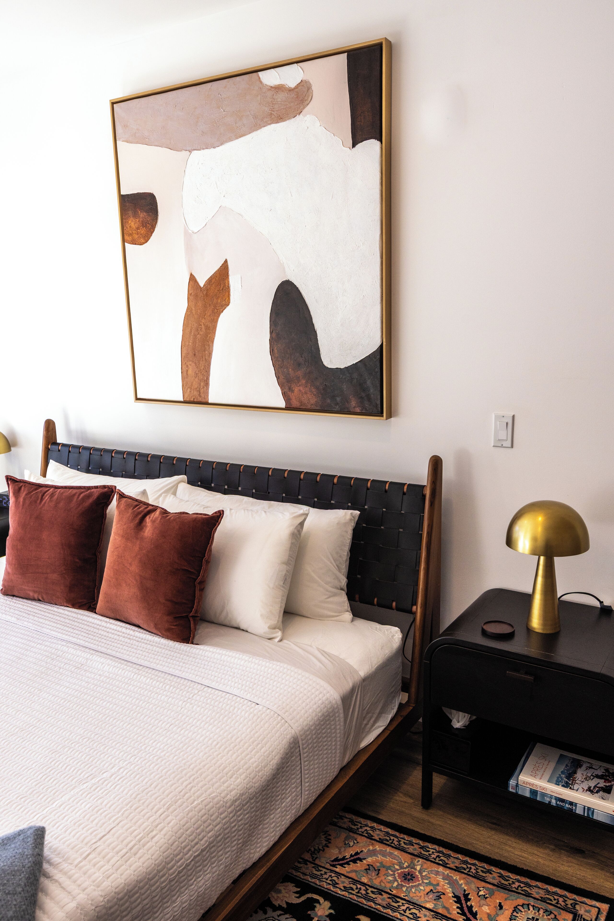 Ein ordentlich eingerichtetes Schlafzimmer mit einem Bett, Kissen, einem Nachttisch mit Lampe und einem an der Wand hängenden abstrakten Gemälde.