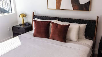 Un lit bien fait, aux couleurs blanches et brunes, avec une peinture abstraite moderne accrochée au-dessus, et une petite table de chevet sur laquelle se trouvent une lampe et une horloge.