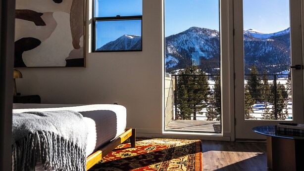 A bedroom with a bed, a large window, a view of snow-covered mountains, and a balcony with a railing.